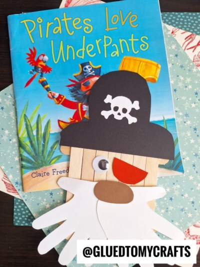 A children’s book titled Pirates Love Underpants sits behind a Popsicle Stick Pirate craft made of paper and craft sticks, featuring a black hat with a skull and crossbones, a white beard, and an orange mouth.