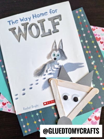 A children’s book titled The Way Home for Wolf rests on colorful paper. Next to it is a popsicle stick wolf face craft made with a popsicle stick triangle, googly eyes, and paper ears. The handle reads @GLUEDTOMYCRAFTS. This adorable setup pairs perfectly with other storytime favorites like a three little pigs craft or even a playful popsicle stick pig creation!.