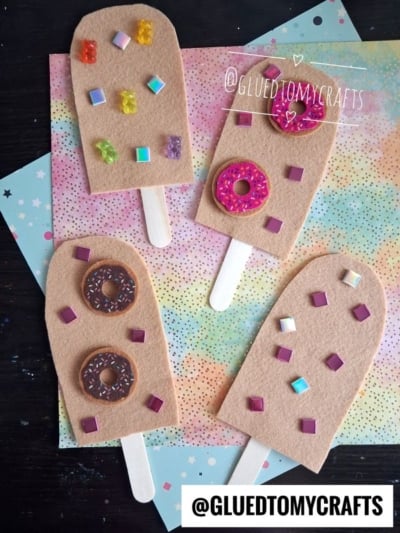 Four felt pretend play popsicles with decorative toppings and wooden sticks are placed on pastel-colored star and rainbow patterned paper. The popsicles are decorated with felt squares and painted donut shapes.