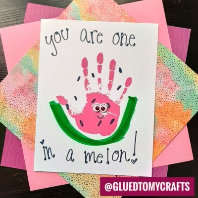 A handmade handprint watermelon card featuring a pink handprint decorated as a watermelon slice with googly eyes and painted seeds. The text reads "You are one in a melon!" on colorful patterned paper.