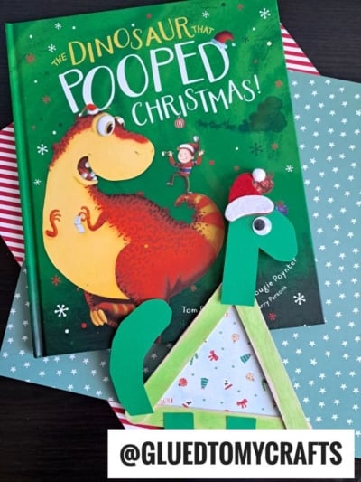 A children’s book titled The Dinosaur That Pooped Christmas sits beside a Popsicle Stick Dinosaur craft, made from paper and popsicle sticks and decorated with a Santa hat. A label reads @GLUEDTOMYCRAFTS.