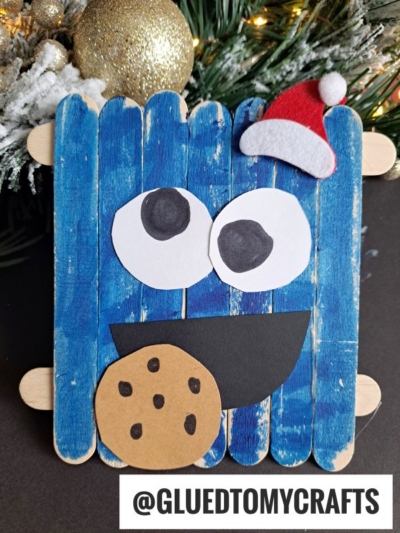 A popsicle stick Cookie Monster craft features big googly eyes, a black mouth clutching a paper cookie, and a tiny red Santa hat. Snowy pine branches and gold ornaments decorate the background.