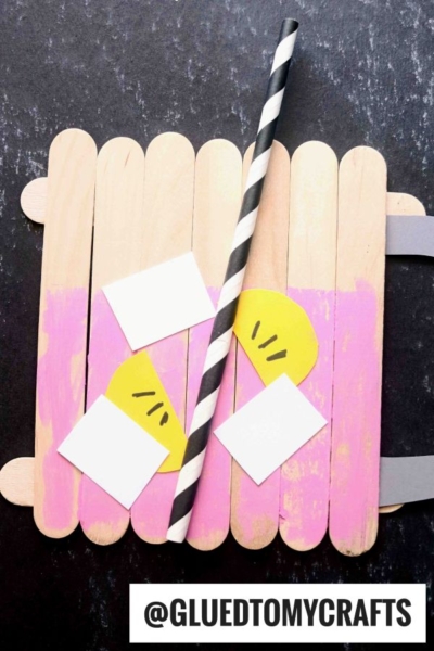 A popsicle stick pink lemonade craft painted partly pink, decorated with yellow and white paper, a black-and-white striped straw, and a handle shaped like a lemonade pitcher. Text reads @GLUEDTOMYCRAFTS.