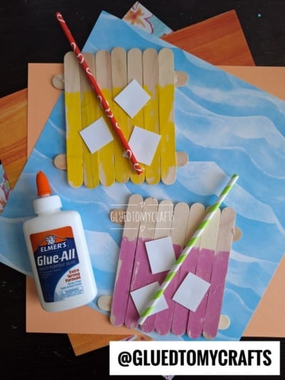 Three popsicle stick rafts, inspired by pink lemonade, are painted yellow and pink and decorated with paper squares and straws. Glue, colored papers, and the handle @GLUEDTOMYCRAFTS are also shown on a table.