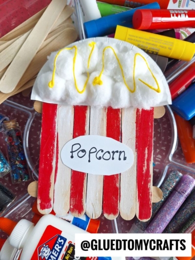 A Popsicle Stick Popcorn project featuring red and white painted sticks, cotton balls with yellow paint for butter, labeled Popcorn, surrounded by craft supplies—a fun and creative craft stick popcorn craft.