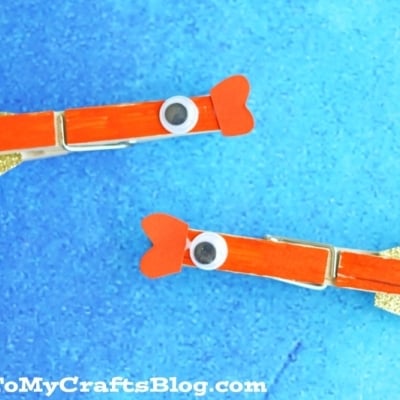 Two clothespin goldfish, decorated with orange paint, googly eyes, red paper fins, and gold glittery tails, are displayed on a blue background. The image features the text GluedToMyCraftsBlog.com at the bottom.