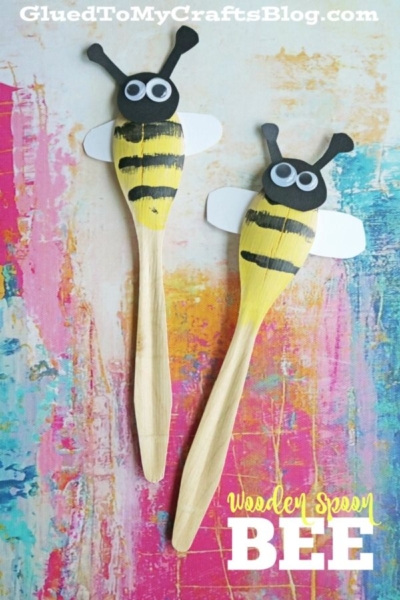 Two wooden spoons painted as bees with black and yellow stripes, black heads, white wings, googly eyes, and black pipe cleaner antennae create an adorable wooden spoon bee craft, placed on a colorful abstract background.
