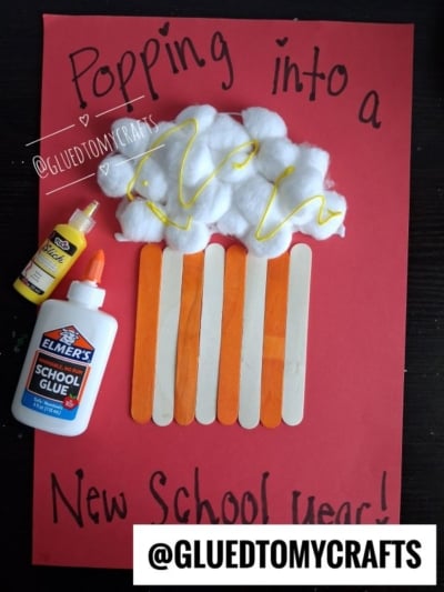 A red paper displays a Popsicle Stick Popcorn-themed craft using cotton balls, popsicle sticks, yellow and white glue, with the text “Popping into a New School Year!” written around the craft.