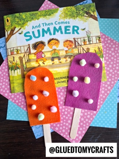 Two colorful felt pretend play popsicles decorated with pom-poms and wooden sticks are placed in front of the children’s book “And Then Comes Summer,” on top of patterned paper. @GLUEDTOMYCRAFTS is written at the bottom.