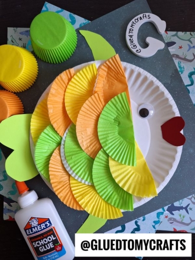 A colorful cupcake liner fish craft made from a paper plate, orange, yellow, and green cupcake liners, with a red mouth and googly eye, lies on a blue background with craft supplies and @GLUEDTOMYCRAFTS label.