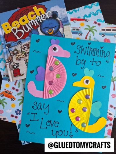 A handmade card with pink and yellow cupcake liner seahorse designs, decorated with gems on blue paper, reads Swimming by to say I Love You! It is placed atop beach-themed books and paper. @GLUETOMYCRAFTS is visible at the bottom.