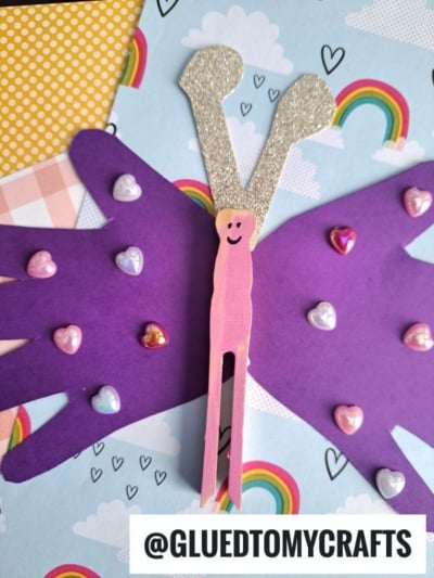 A handprint clothespin butterfly craft made from purple paper handprints for wings, a pink clothespin body with a smiley face, glittery silver antennae, and heart-shaped stickers, displayed on rainbow-patterned paper. Text: @GLUEDTOMYCRAFTS.