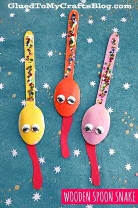 Three colorful Wooden Spoon Snakes—yellow, orange, and pink—decorated with googly eyes, painted tongues, and glitter, are arranged on a blue background with white star patterns.