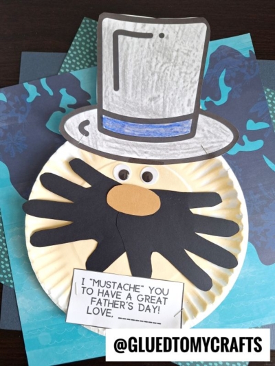 A handmade Father’s Day mustache you card features a paper plate with a large black handprint mustache, brown nose, and paper top hat. A note says, “I ‘MUSTACHE’ YOU TO HAVE A GREAT FATHER’S DAY! LOVE, _____.” @GLUEDTOMYCRAFTS at the bottom.