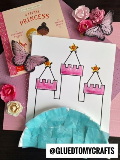 A pink paper plate princess castle drawing with tissue paper at the bottom, surrounded by a book titled A Little Princess, fake flowers, butterfly cutouts, and a @GLUEDTOMYCRAFTS label on a pink polka dot surface.