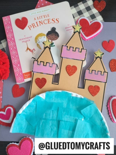 A children’s book titled A Little Princess sits next to a pink paper plate princess castle craft decorated with red hearts, blue tissue paper, and surrounded by heart-shaped felt and paper cutouts on a gray background.