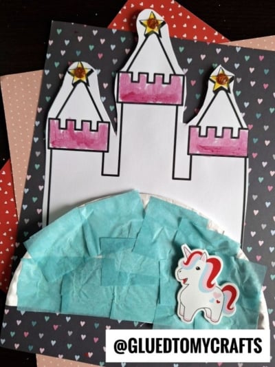 A charming paper plate princess castle scene features a white and pink castle topped with gold stars, blue tissue paper for water or sky, and a small unicorn cutout, all set on heart-patterned paper. @GLUEDTOMYCRAFTS is written at the bottom.