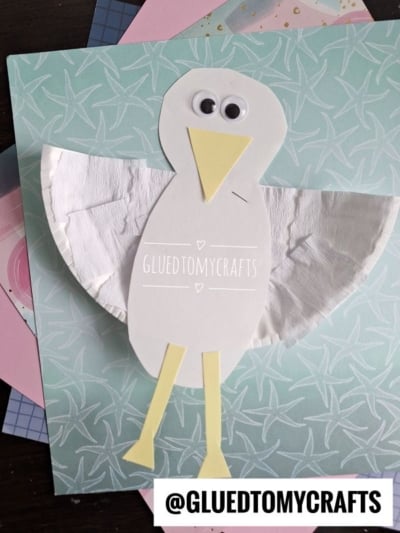 A handmade paper plate seagull craft made from white paper and a paper plate, with googly eyes, a yellow paper beak and legs, displayed on a pastel patterned background. The text @GLUEDTOMYCRAFTS appears at the bottom.