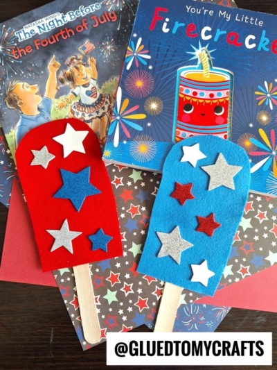 Two handmade felt pretend play popsicles with stars, one red and one blue, on wooden sticks. Behind them are colorful Fourth of July-themed children’s books and patterned paper. @GLUEDTOMYCRAFTS is written at the bottom.