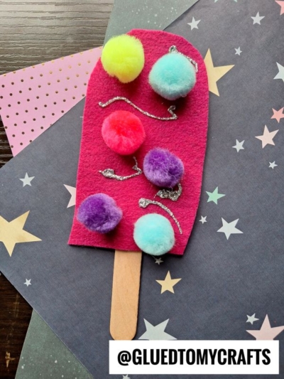 A popsicle-shaped craft made from red felt, decorated with colorful pom-poms and silver glitter on a wooden stick, is perfect for felt pretend play popsicles and looks charming on star-patterned paper.
