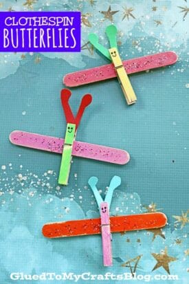 Three colorful clothespin butterfly crafts with glittery wings and smiling faces are displayed on a teal, star-patterned background. The text reads Clothespin Butterflies and GluedToMyCraftsBlog.com.