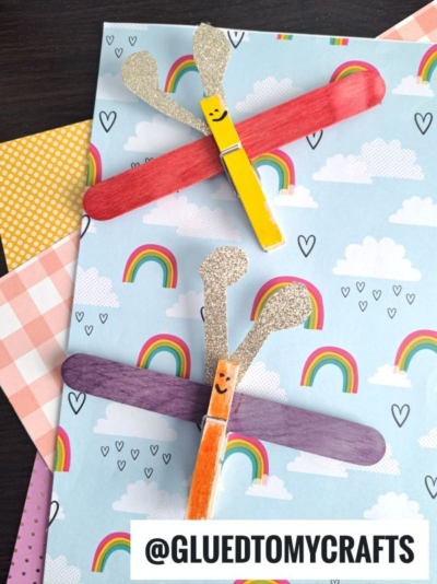 Two charming clothespin crafts—a kangaroo and a clothespin butterfly, both with glittery accents—are attached to colored popsicle sticks, displayed on rainbow-themed paper with a “@GLUEDTOMYCRAFTS” label below.