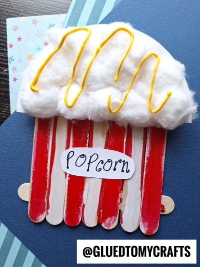 A Popsicle Stick Popcorn craft made from red and white painted sticks, topped with cotton balls as popcorn and yellow squiggles as butter, sits on blue paper. A Popcorn label is attached. Handle: @GLUEDTOMYCRAFTS.