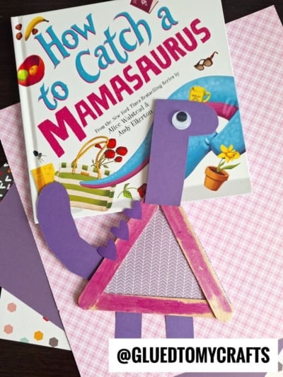 A childrens book titled How to Catch a Mamasaurus lies on patterned paper next to a purple craft stick dinosaur made from construction paper and craft sticks. The username @GLUEDTOMYCRAFTS appears at the bottom.