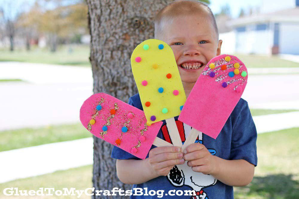 Smiling child holds three colorful felt pretend play popsicles crafted from popsicle sticks, decorated with pom-poms and glitter, standing outdoors by a tree.