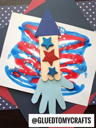 A handmade rocket craft made from Popsicle Stick Fireworks, paper stars, and a blue paper handprint sits on abstract red, white, and blue art, surrounded by star-patterned paper. @GLUEDTOMYCRAFTS is written at the bottom.