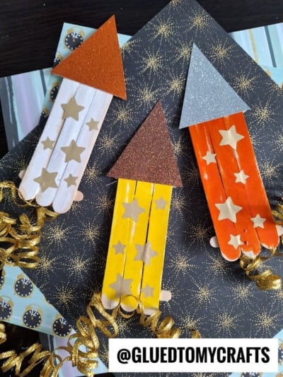 Three colorful Popsicle Stick Fireworks with glittery tops and gold star stickers are displayed on a dark background with star patterns. Gold curly ribbons decorate each craft stick firework. @GLUEDTOMYCRAFTS is written at the bottom.