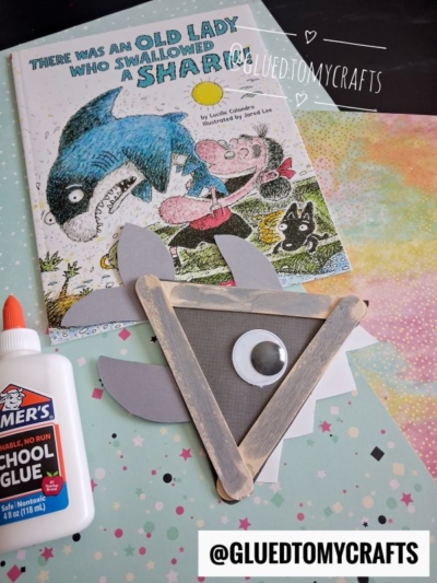 A children’s book, a popsicle stick shark craft made of gray triangles and a googly eye, white glue bottle, and @gluedtomycrafts handle on a colorful, speckled surface.