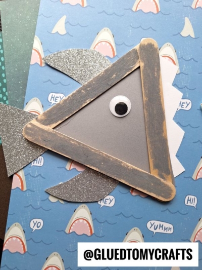 A popsicle stick shark craft made from gray-painted sticks, a googly eye, and glittery fins sits on shark-themed paper. The username @GLUEDTOMYCRAFTS appears at the bottom—perfect for any shark kid craft project.