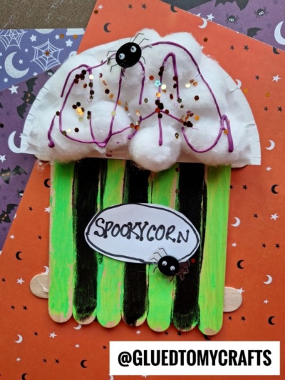 A Halloween Popsicle Stick Popcorn craft made with neon green and black sticks, cotton ball and purple string popcorn, googly-eyed spiders, glitter, and a “Spookycorn” sign on an orange background with bats and webs.
