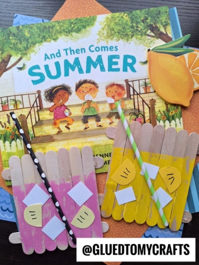 A children's book titled And Then Comes Summer with two popsicle stick pink lemonade crafts—each featuring a paper straw and lemon slices—resting on top of the book.