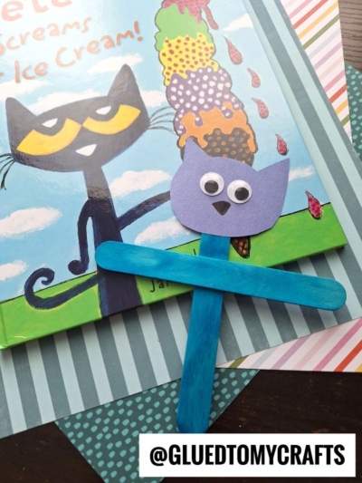 A Pete The Cat Puppet made from a craft stick, featuring googly eyes and blue fur, lies on a striped surface next to the book Pete the Cat: I Scream for Ice Cream! @GLUEDTOMYCRAFTS is written at the bottom.
