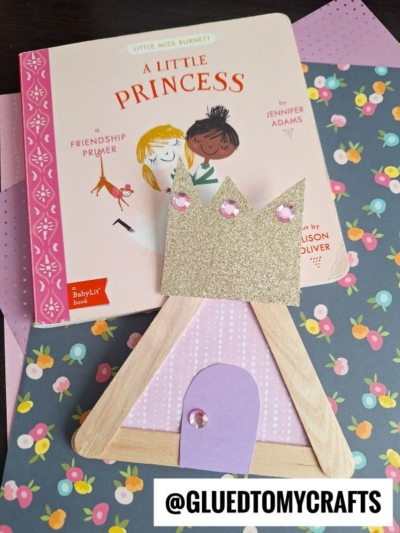 A children’s book titled A Little Princess sits on colorful floral paper next to a Popsicle Stick Princess Castle craft, made with popsicle sticks, glittery paper, and pink gems. @GLUEDTOMYCRAFTS is written at the bottom.