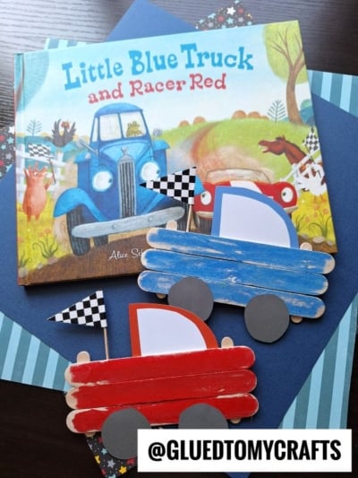 A children’s book titled “Little Blue Truck and Racer Red” is displayed with two craft stick race cars made from red and blue painted popsicle sticks and checkered flags, arranged on blue and striped paper backgrounds.