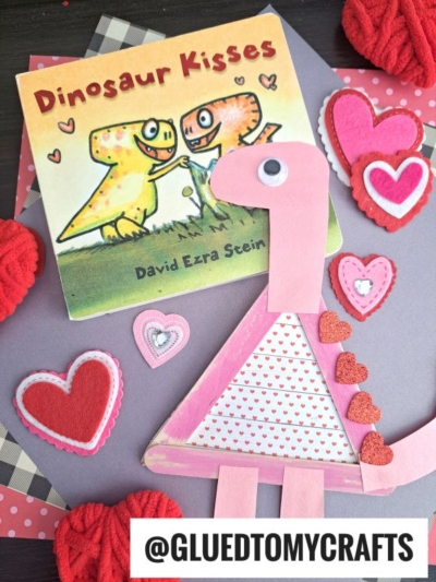 A children’s book titled Dinosaur Kisses sits on a table decorated with pink, red, and white felt hearts. In front is a handmade pink Popsicle Stick Dinosaur craft made with paper and glitter hearts. @GLUEDTOMYCRAFTS is at the bottom.