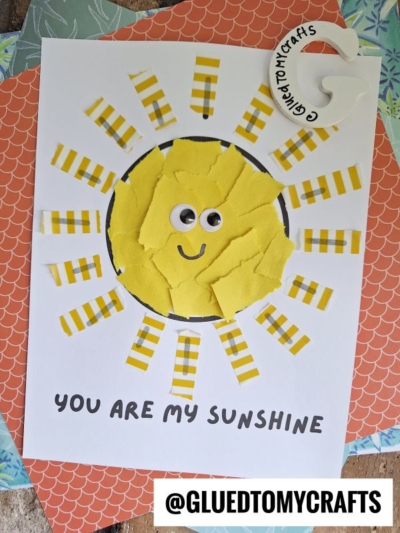 A children’s craft of a sunshine card features a washi tape sun with a yellow, torn-paper center, googly eyes, and rays made from yellow-striped paper. Below, text reads “YOU ARE MY SUNSHINE.” The handle @GLUETOMYCRAFTS is at the bottom.