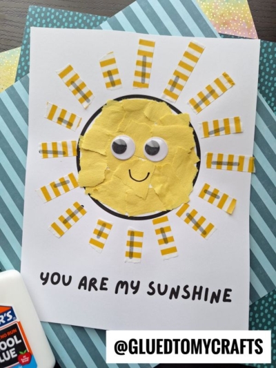 A handmade sunshine card featuring a yellow sun with googly eyes and a smile, surrounded by striped washi tape rays, sits on white paper labeled YOU ARE MY SUNSHINE. A glue bottle rests beside this cheerful craft.