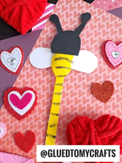 A handmade Valentine’s Day card features a bumble bee wooden spoon crafted from paper and a yellow stick, surrounded by red and pink felt hearts on a patterned background. @GLUEDTOMYCRAFTS is written at the bottom.