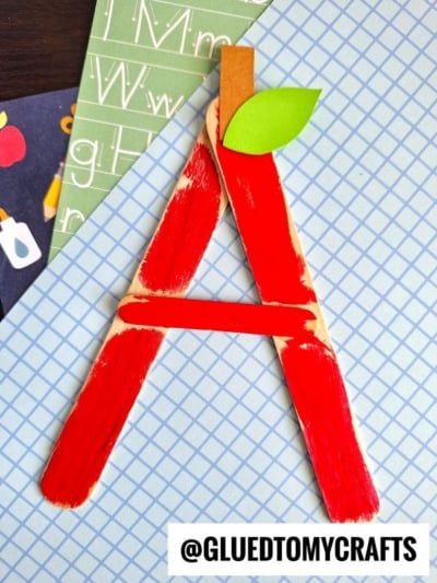 A popsicle stick “A” made from red-painted sticks with a green paper leaf on top, mimicking an apple, is placed on graph paper with school-themed backgrounds. @GLUEDTOMYCRAFTS is printed at the bottom.
