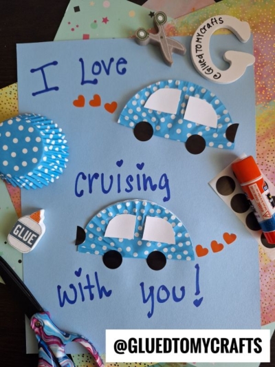 A handmade card says I Love cruising with you! and features two blue cupcake liner cars with paper wheels and hearts. Craft supplies like scissors, glue, and markers are scattered around.