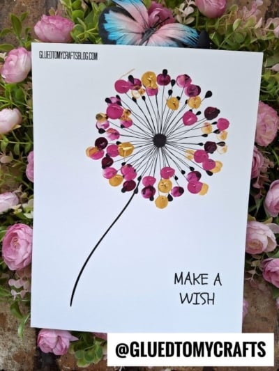 A handmade Thumbprint Dandelion art piece features colorful fingerprint petals on white paper, set atop pink flowers. Text reads “MAKE A WISH” and “@GLUEDTOMYCRAFTS.” A charming dandelion wish craft for all ages!.