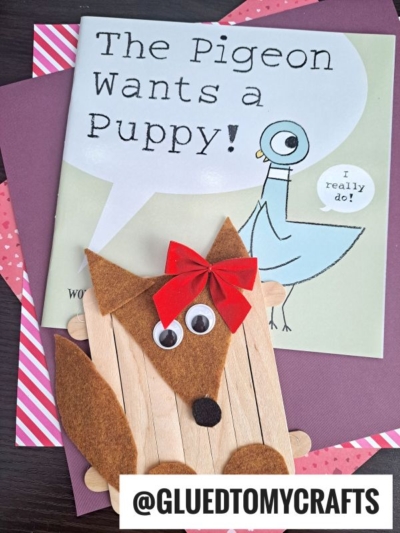 A book titled The Pigeon Wants a Puppy! lies on pink and purple paper. In front is a craft stick dog fox craft made from brown felt, googly eyes, a red bow, and wooden sticks. @GLUEDTOMYCRAFTS is written at the bottom.