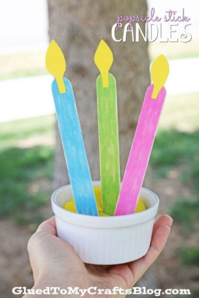 Three colorful Popsicle Stick Candles—blue, green, and pink with yellow flames—are held upright in a white cup, shown outdoors with a tree and grass blurred in the background.