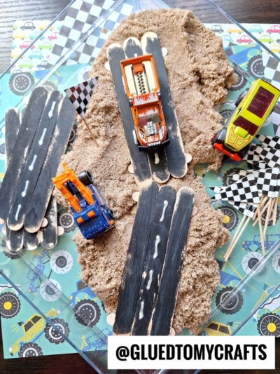 Toy race cars speed along popsicle stick race tracks set over piles of kinetic sand, with checkered flags waving and a monster truck-themed backdrop. The @GLUEDTOMYCRAFTS watermark is at the bottom.