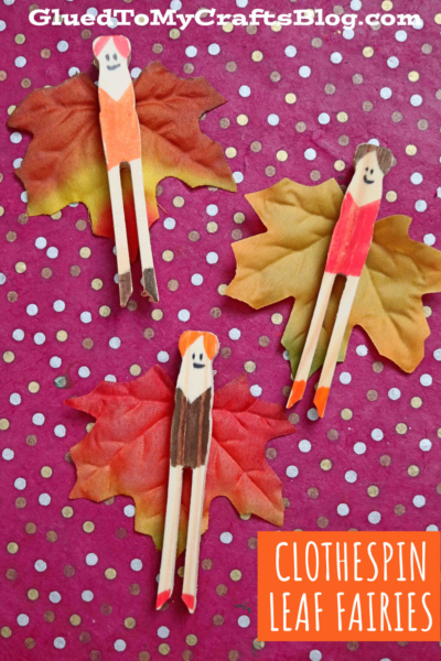 Three handmade fairy clothespin leaf fairies with drawn faces and colorful outfits sit on artificial autumn leaves, arranged on a pink polka dot background. Text reads “Clothespin Leaf Fairies.”.