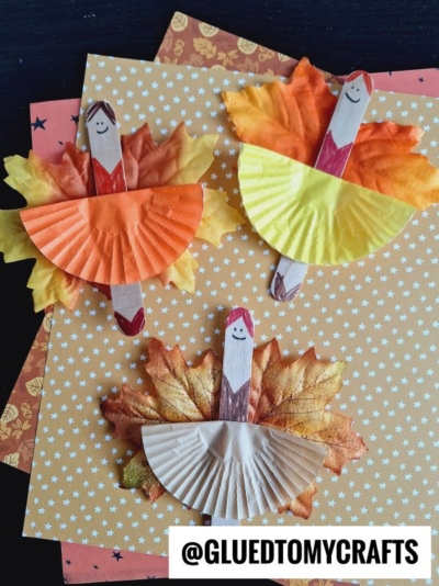 Three craft stick dolls with drawn faces wear cupcake liner dresses and faux leaf wings, transforming them into whimsical leaf fairy ballerinas. The dolls are placed on patterned paper with small stars. @GLUEDTOMYCRAFTS is written at the bottom.